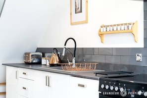 Eine moderne Küche mit Wasserkocher, Toaster und Herd. Es ist ein Waschbecken mit Wasserhahn, ein Geschirrständer und ein Regal mit Gläsern vorhanden. Die Küche hat weiße Schränke und eine graue Spritzschutzwand.