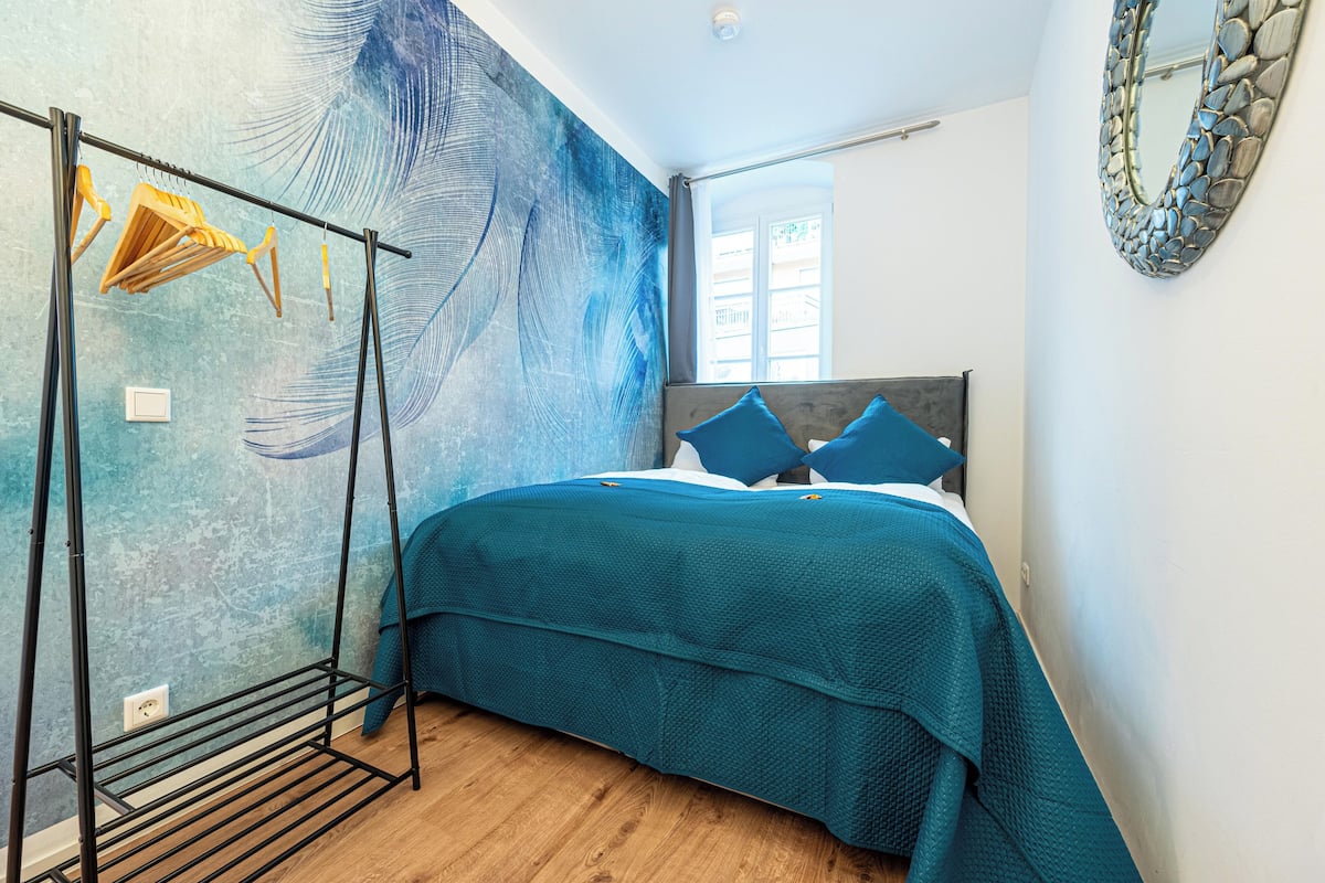 Ein Schlafzimmer mit einem Bett, einem Wäscheständer, einem großen abstrakten Wandbild, einem Fenster und einem spiegelnden Wandbild mit Steinrahmen.