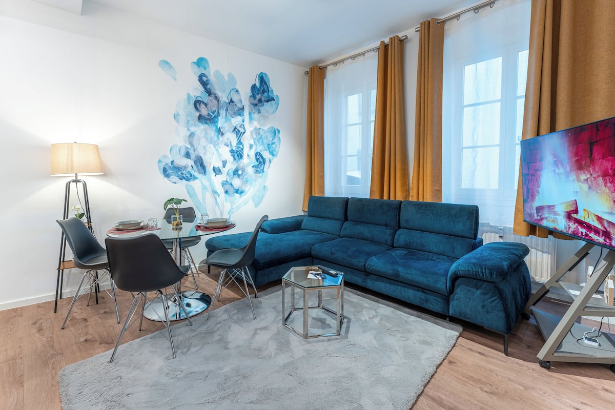 Ein modernes Wohnzimmer mit einem blauen Sofa, einem Glastisch, einem Essbereich mit Stühlen und einem großen abstrakten Gemälde.