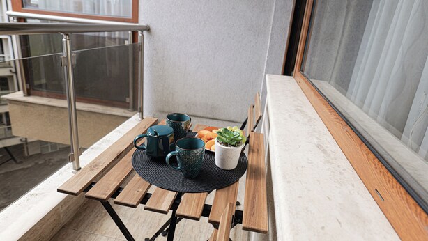 A balcony with a wooden table, two green mugs, a white pot with a plant, and a metal railing. - SouthPark Luxe - Free Underground Parking (Sofia)
