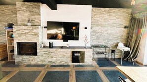Een moderne woonkamer met een flatscreen televisie aan een stenen muur, een open haard en een eethoek met een tafel en stoelen.