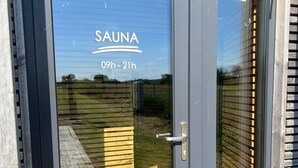 Een glazen deur met de tekst 'SAUNA 09h - 21h', een metalen handvat en een reflectie van een houten terras.