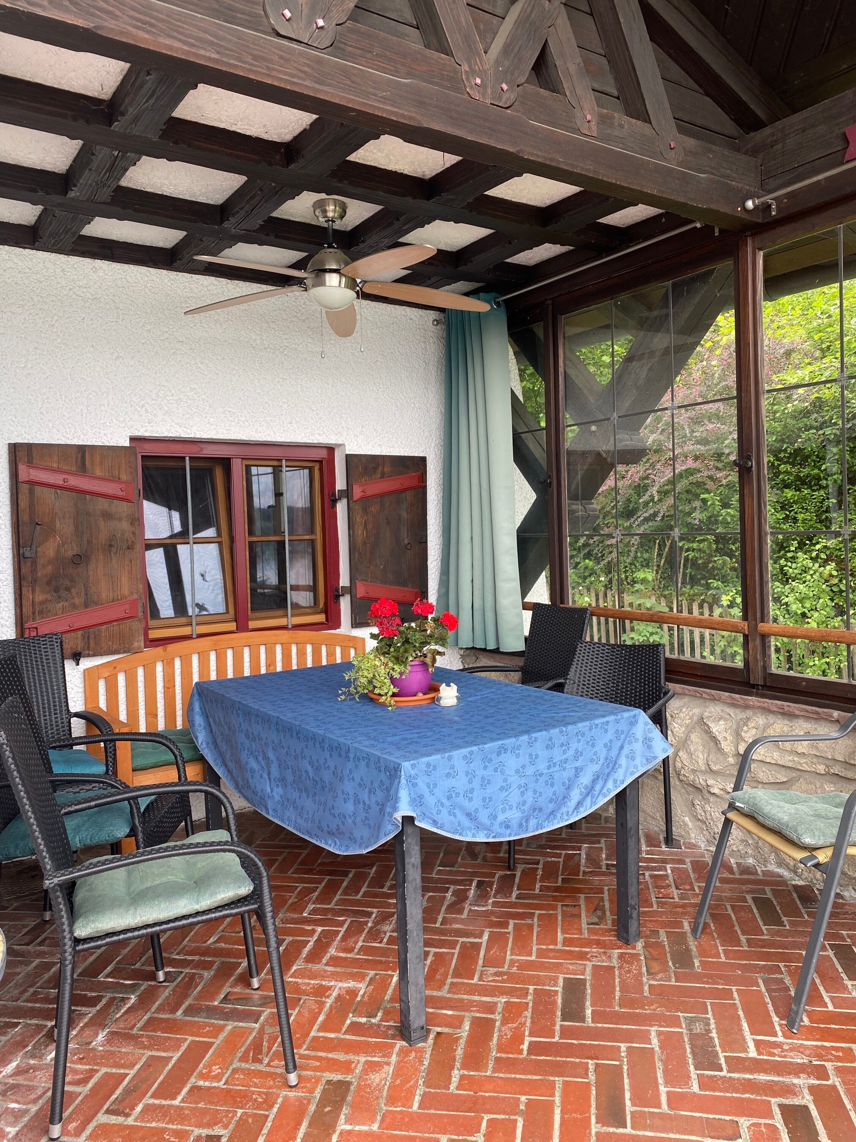 Eine überdachte Terrasse mit Tisch und Stühlen, einem Deckenventilator und Blick auf Grünflächen.