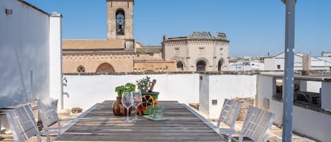 Un patio al aire libre con una mesa y sillas, vista de un edificio histórico con un campanario, y un cielo azul despejado.