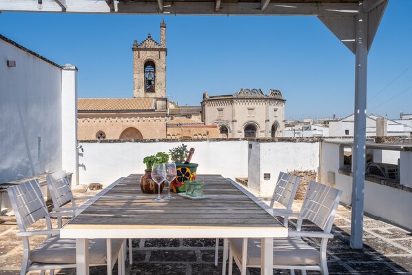 Un patio al aire libre con una mesa y sillas, vista de un edificio histórico con un campanario, y un cielo azul despejado.