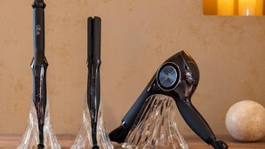 Trois appareils de coiffure sont posés sur une surface en bois, devant un mur texturé, et un objet rond se trouve à l’arrière-plan.