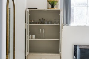 Ein weißes Schranksystem mit Regalen, auf denen Glaswaren stehen, und einem spiegelnden Wandbild mit goldener Rahmung.