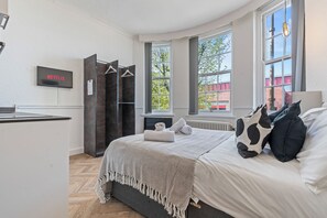 Ein Schlafzimmer mit einem Bett, einem Fernseher, der 'NETFLIX' zeigt, einem Heizkörper und einem Fenster mit Blick auf Bäume.