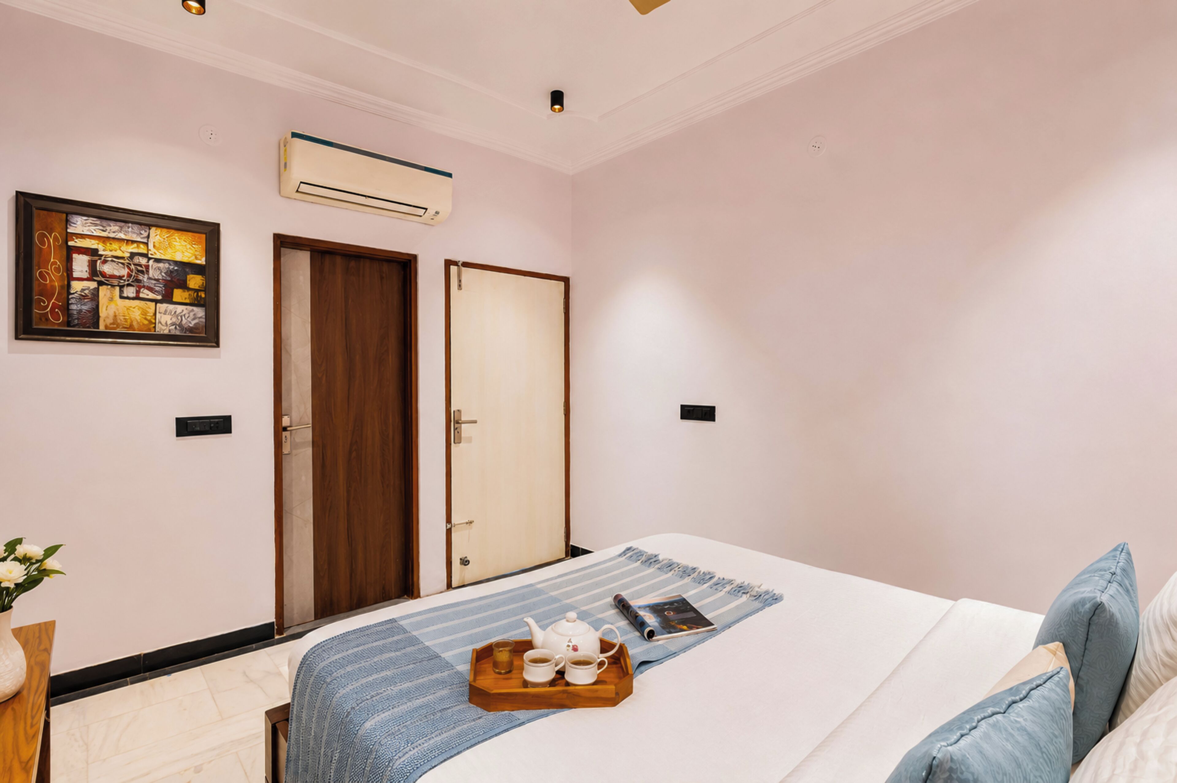 A hotel room with a bed, a bedside table with a tray of tea, a framed picture, and a door.