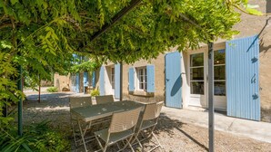 A covered outdoor seating area with a table and chairs, surrounded by trees and a building with blue shutters. - Holiday Home "Mas des Amandiers" with Private Pool, Terrace, and Wi-Fi (Jonquières)