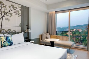 A hotel room with a bed, a sofa, a small table, and a large window offering a view of the landscape. - Amoravida by 7 Apple Resorts Khandala (Lonavala)