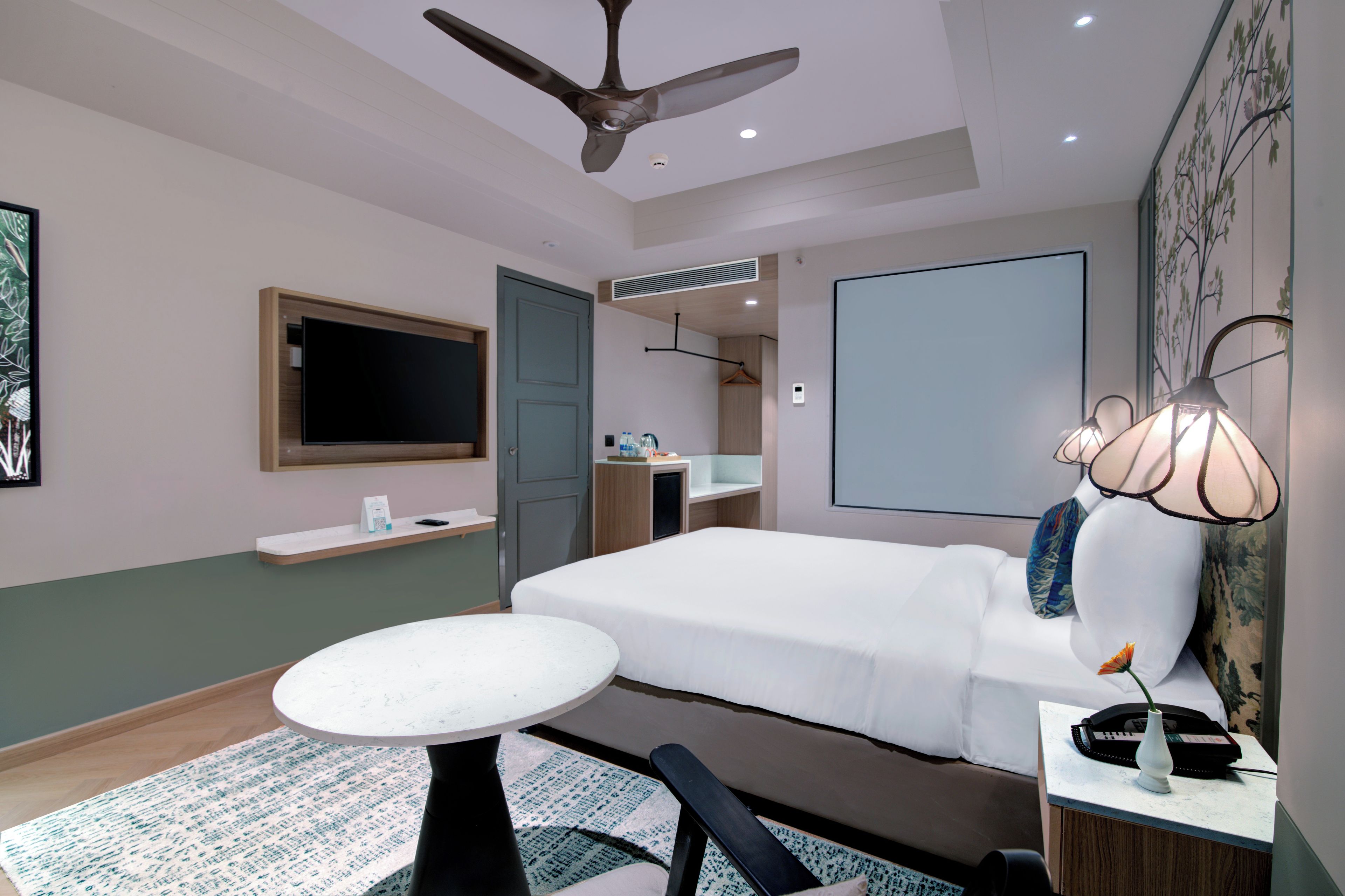 A hotel room with a bed, a desk with a lamp, a TV, a ceiling fan, and a bathroom with a mirror and towel rack.