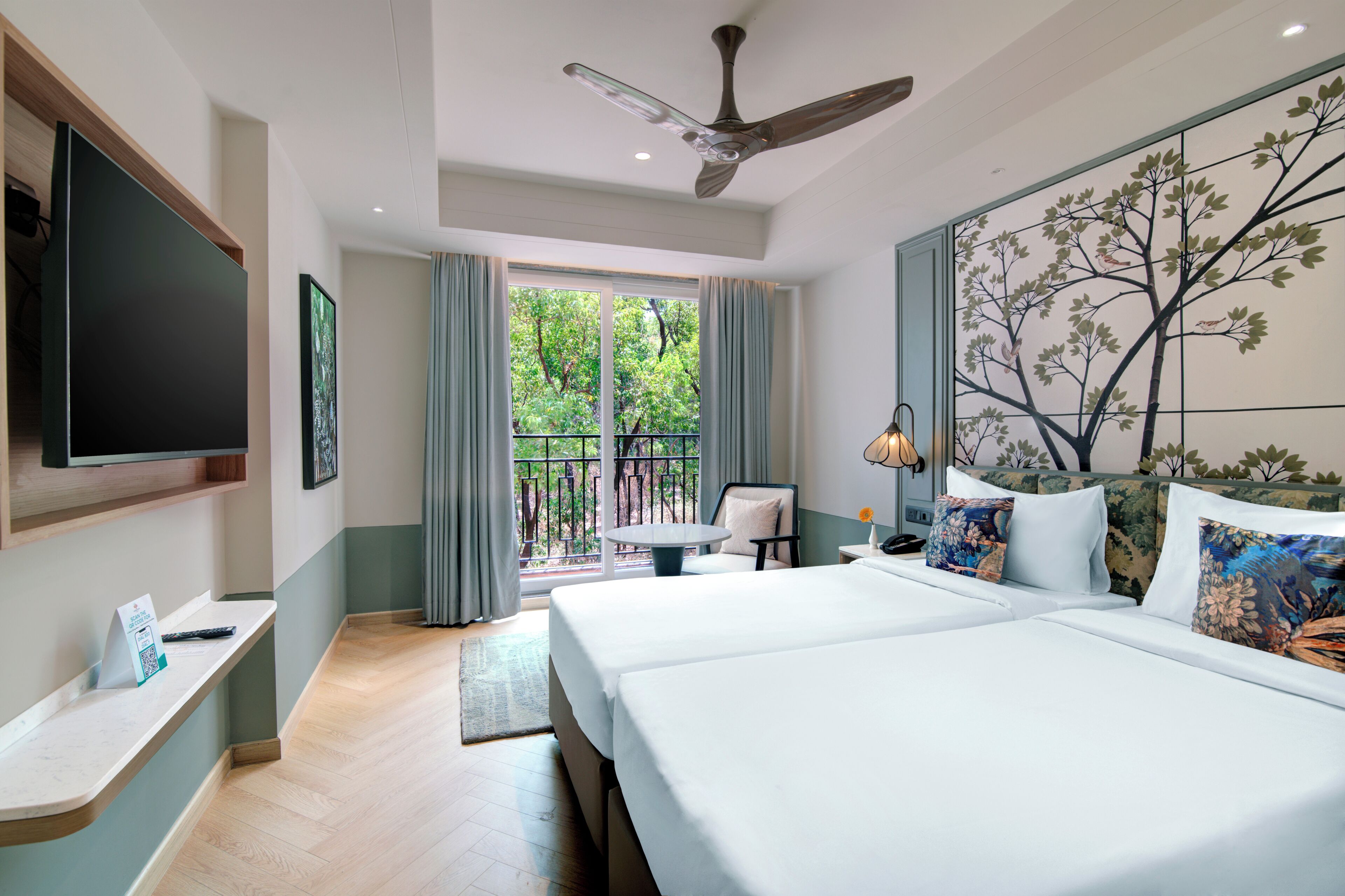 Habitación de hotel con cama, escritorio, silla, televisor y balcón con vista a árboles.