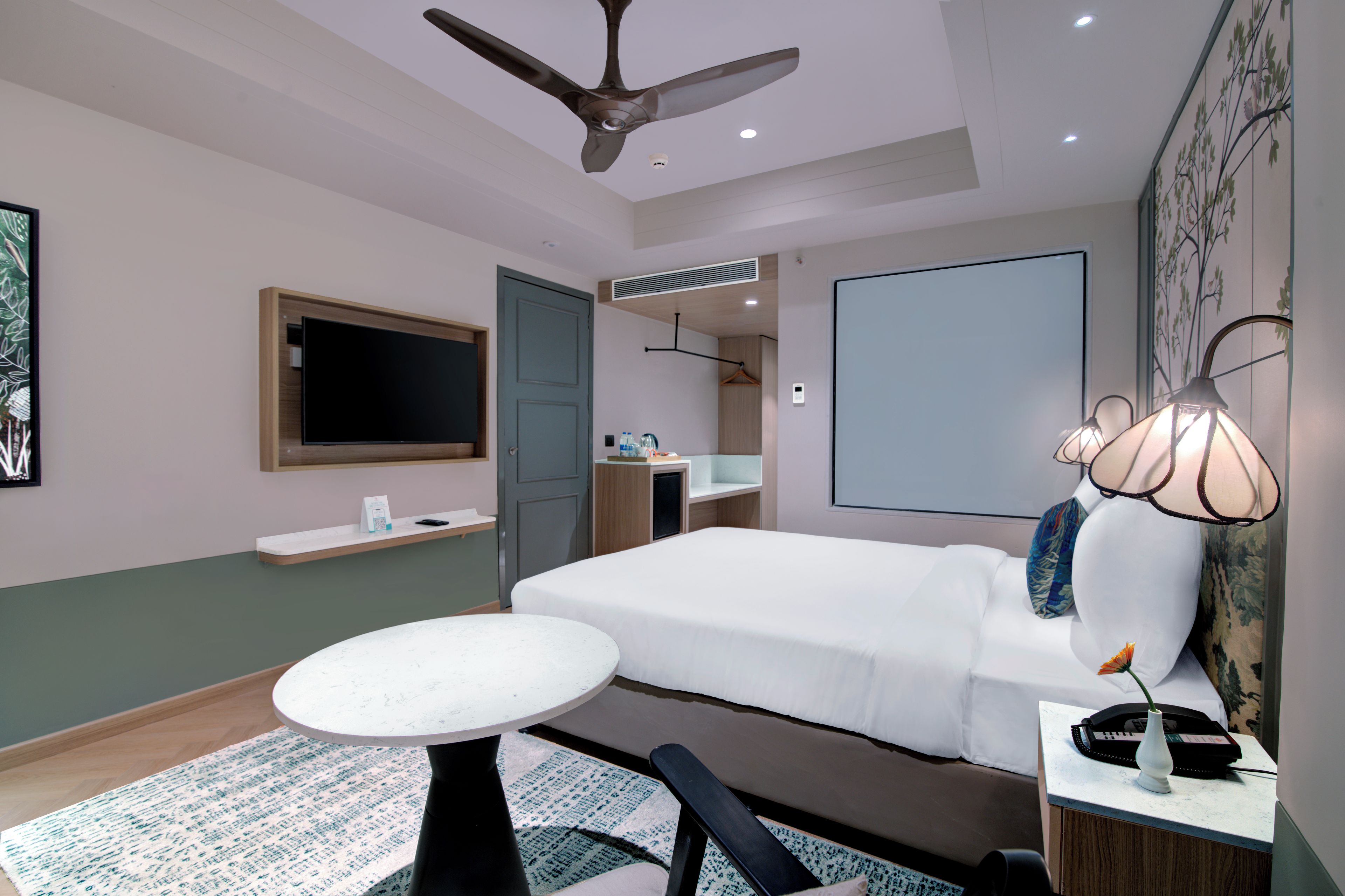 Quarto de hotel com uma cama, uma escrivaninha com luminária, televisão, ventoinha no teto e um banheiro visível através de uma porta aberta.