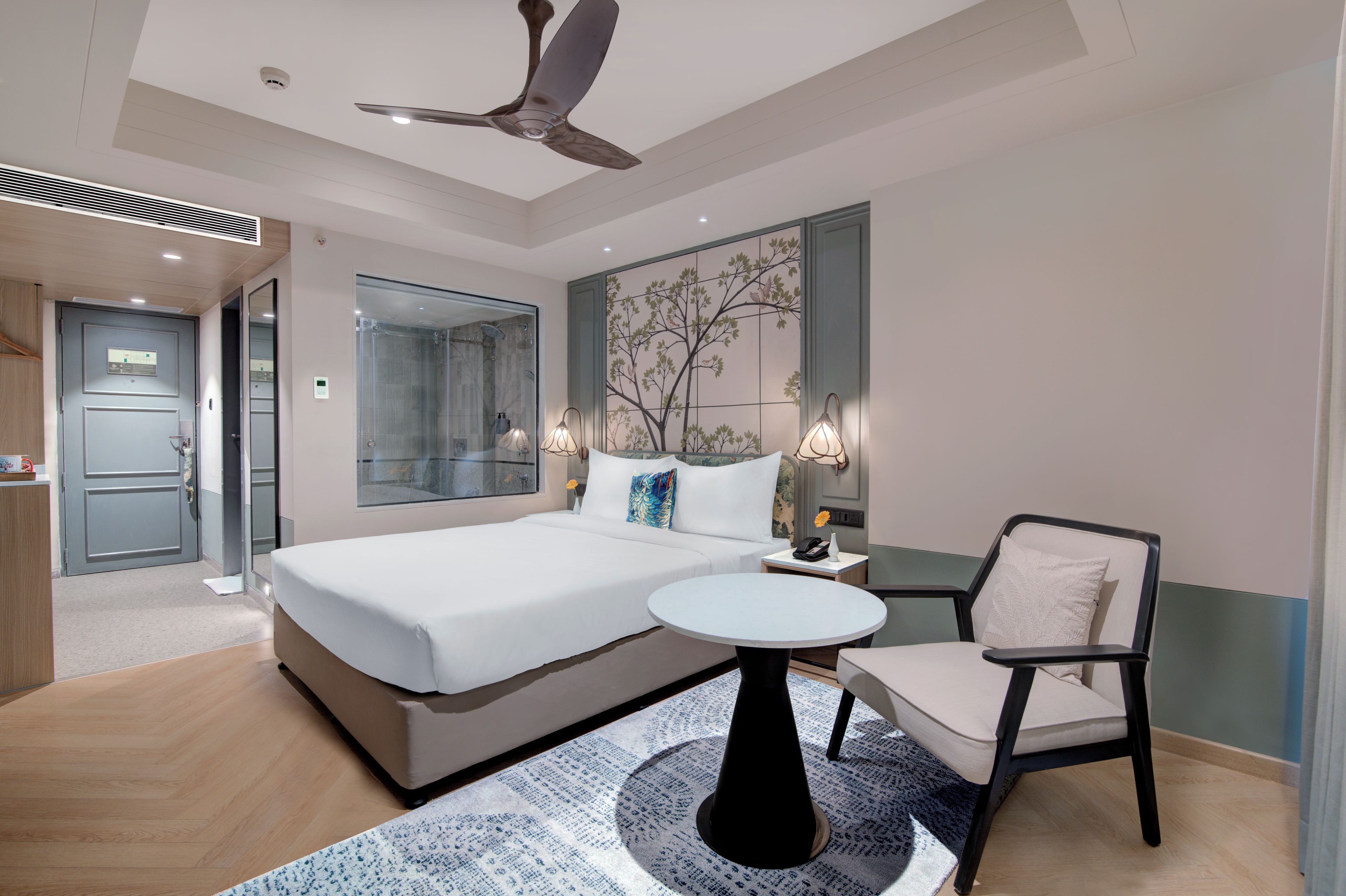 Habitación de hotel moderna con cama, una silla, una mesita y ventilador de techo, además de ducha.
