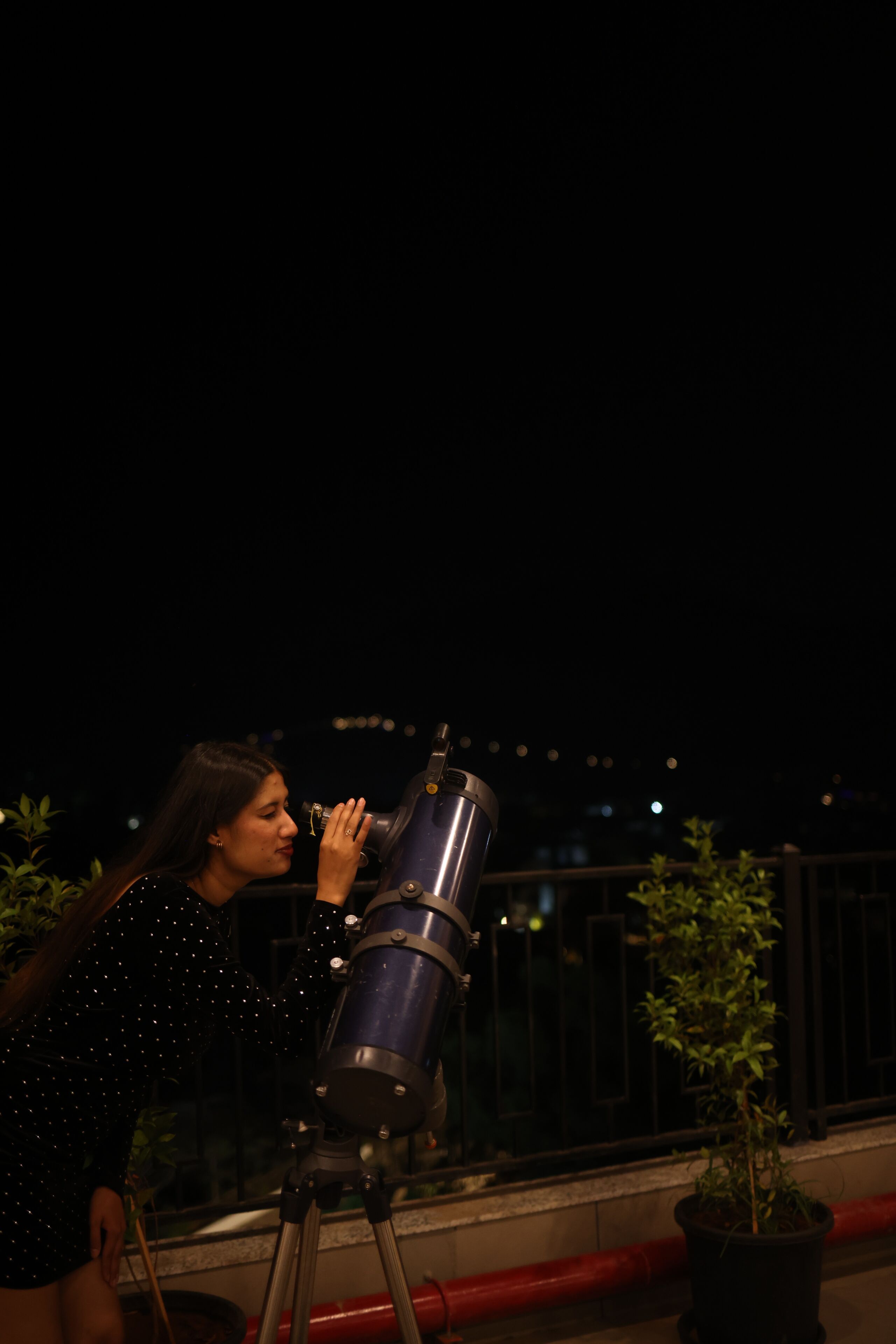 A person using a telescope on a balcony with potted plants and a railing.