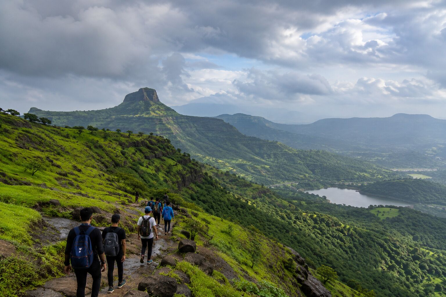 A group of hikers on a rocky trail with a mountainous landscape and a lake in the distance.