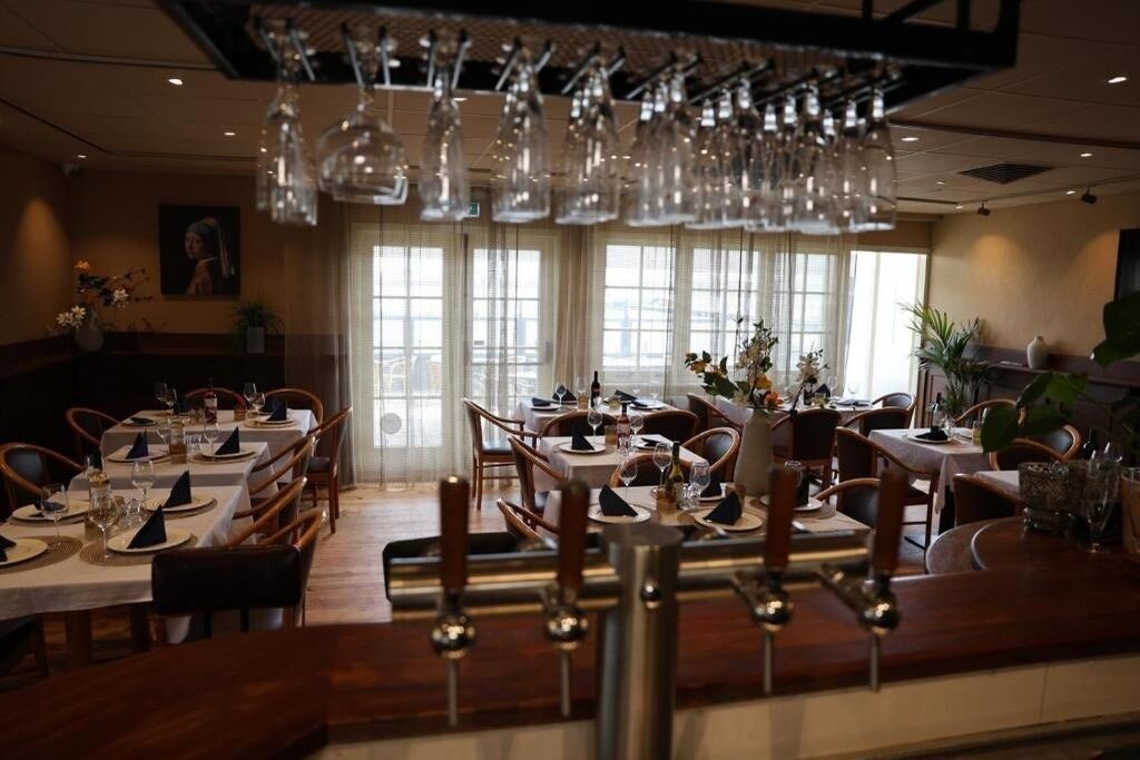 Een restaurant met meerdere eettafels, hangende lampen en grote ramen met gordijnen.