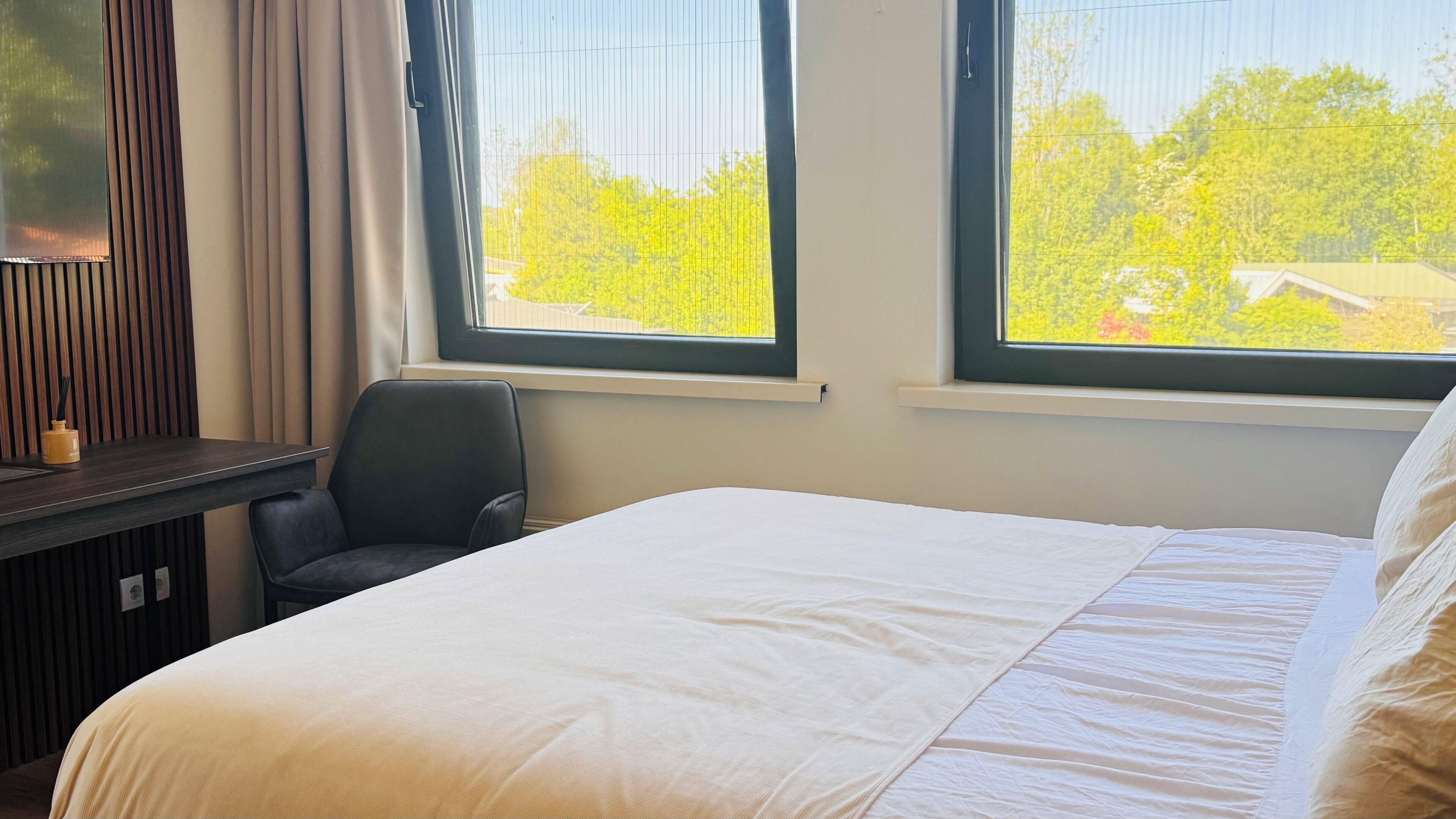 Quarto de hotel com uma cama, uma escrivaninha, uma cadeira e duas janelas com vista para árvores e edifícios.