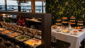 Mesa de buffet con diferentes alimentos, un mantel blanco y vista al agua.