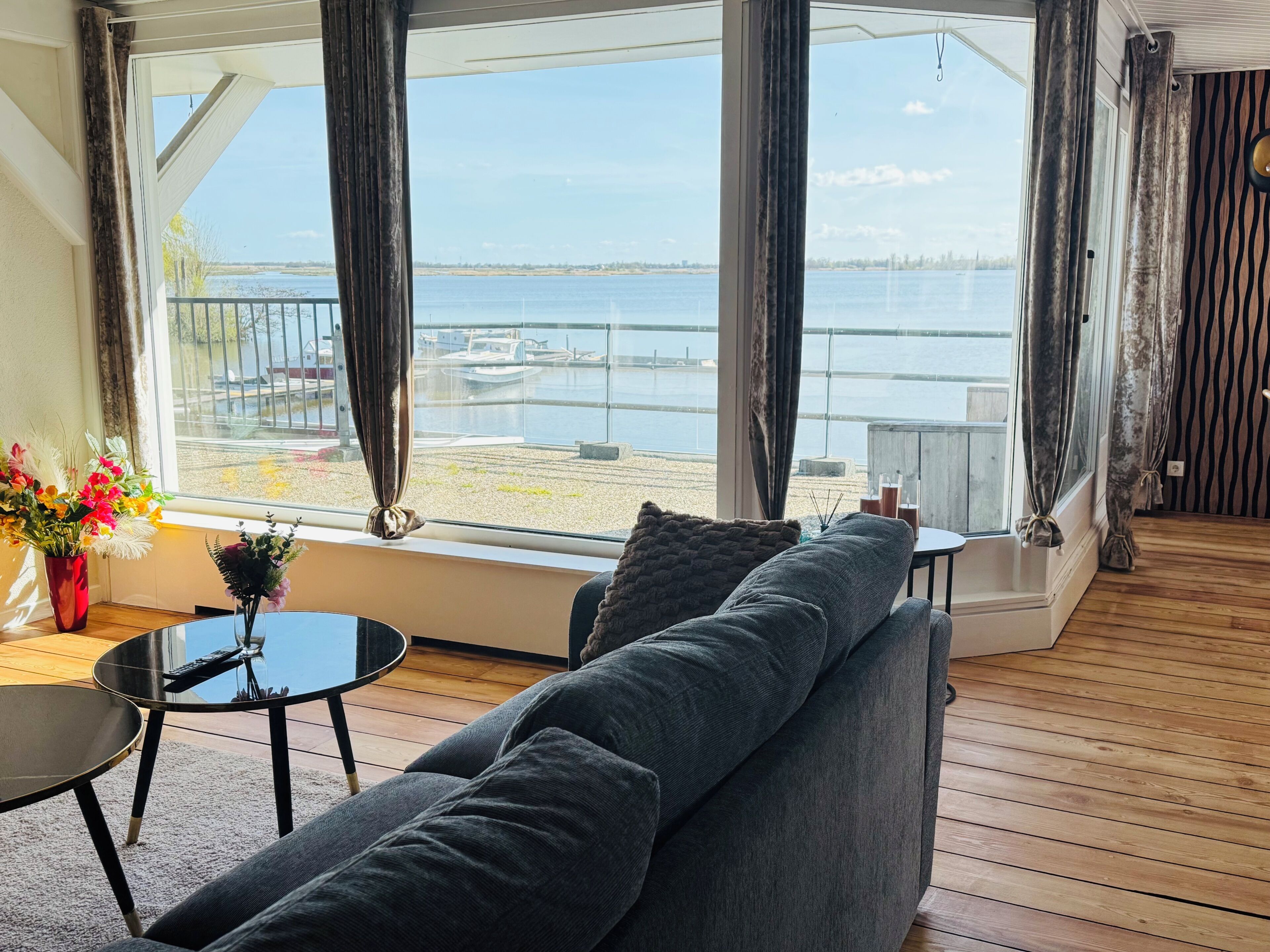 A room with a large window, a grey sofa, a small round table, and a vase of flowers.