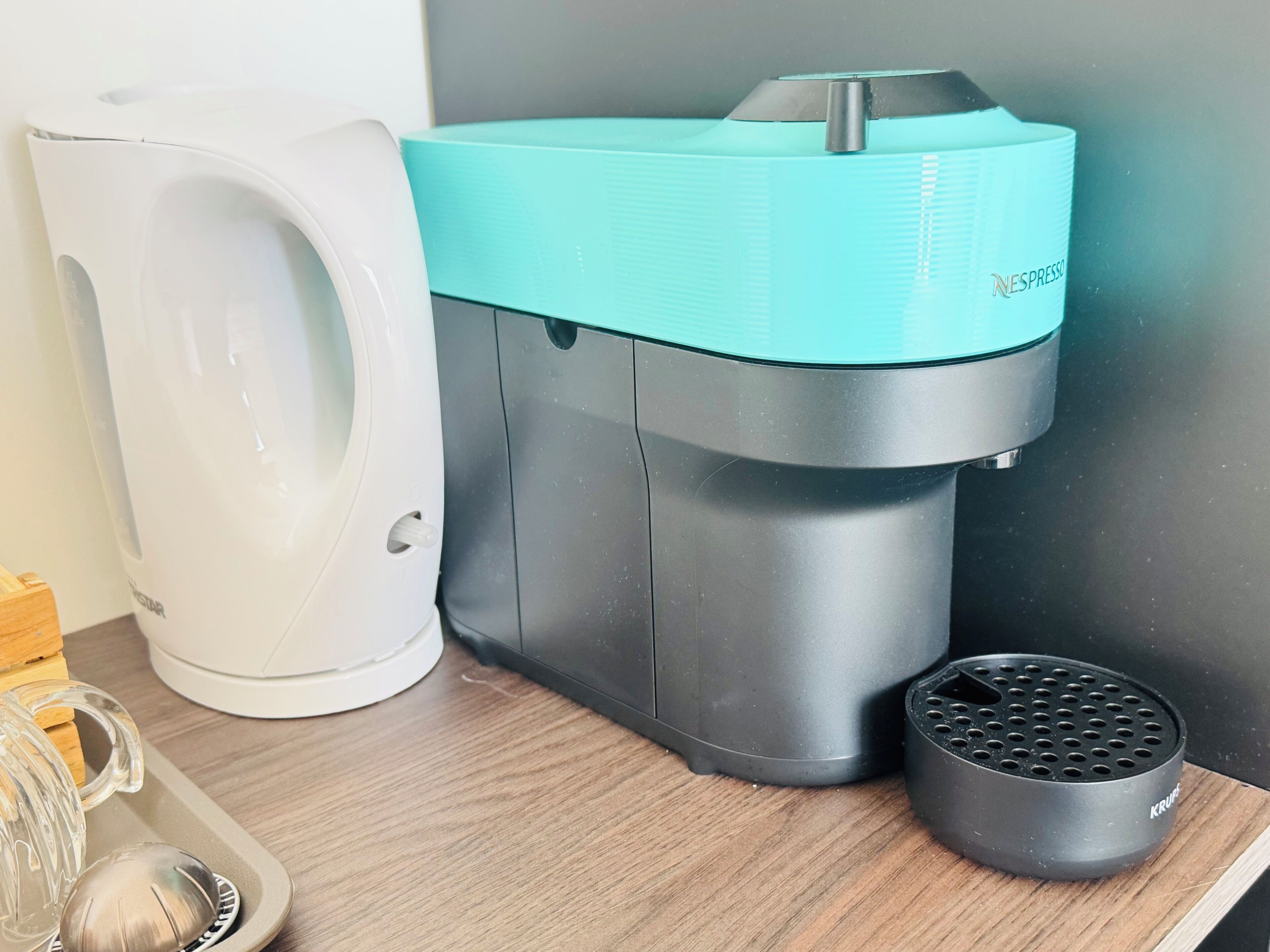 A coffee maker with a teal top and a white base, a black coffee pot, and a small black device on a wooden surface.