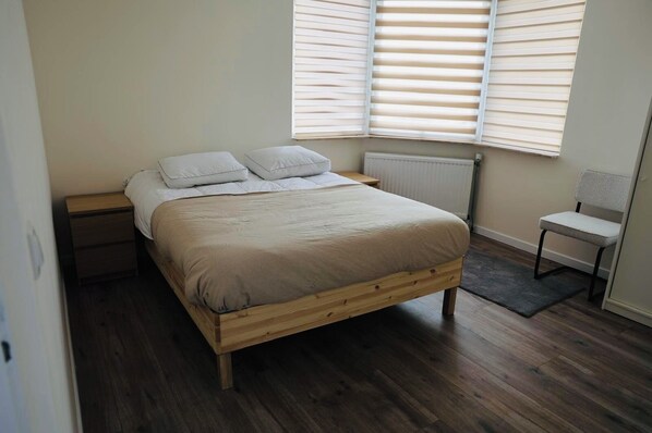 A bedroom with a wooden bed, white bedding, a nightstand, a radiator, a chair, and a window with blinds. - Weromerie Premium Hotel and Apartment (Wormer)