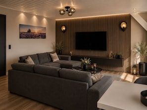 Un salon moderne avec un canapé d’angle, un téléviseur à écran plat et une étagère murale agrémentée d’objets décoratifs.