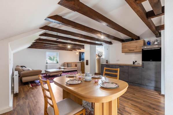 A dining area with a wooden table set for four, a kitchen with modern appliances, and exposed wooden beams on the ceiling.