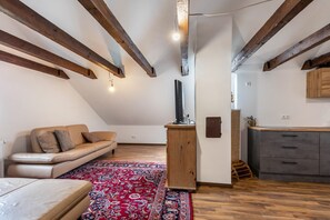 A loft-style room with exposed wooden beams, a sofa, a TV on a wooden stand, a kitchen area, and a patterned rug.