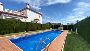 Una piscina rectangular con un deck de madera, rodeada por un seto cuidado y un edificio blanco con techo de tejas.