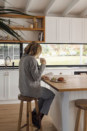 A person sitting at a kitchen island with a window view, white cabinetry, and a wooden stool. - Relaxed Coastal Retreat in Falcon – Perfect for Families (Falcon)