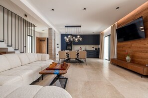 A modern living room with a sofa, a coffee table with chess pieces, a dining area with chairs, a kitchen with a TV, and a staircase. - Villa Krišto Zadarvillas (Privlaka)