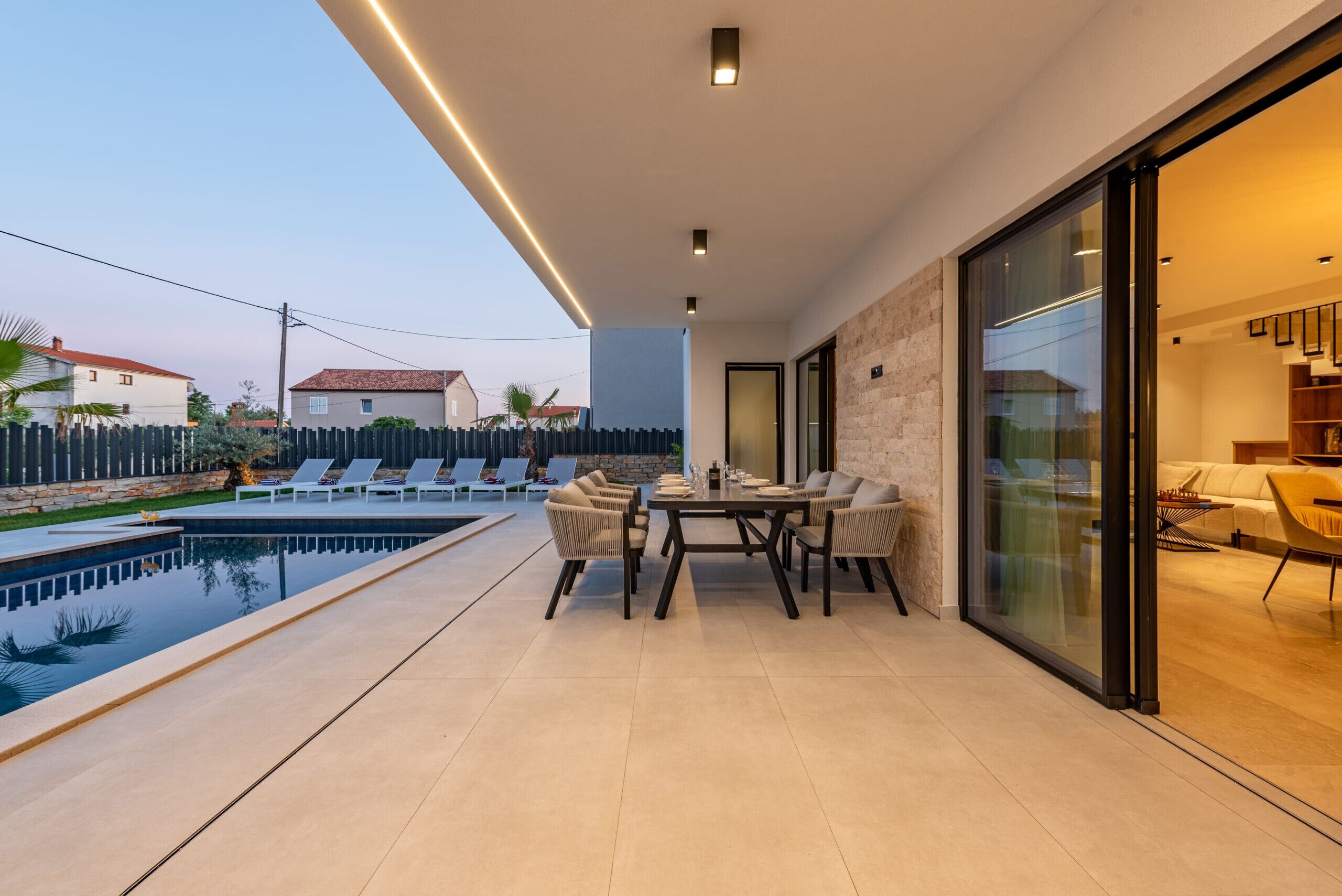A modern outdoor patio with a pool, a dining table with chairs, and a glass door leading to an interior space.