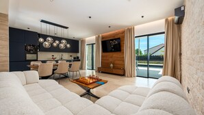 Een moderne woonkamer met een grote bank, een eethoek met stoelen, een keuken met donkere kastjes, en een televisie bevestigd aan een houten muur.