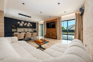 A modern living room with a large sofa, a dining area with chairs, a kitchen with dark cabinets, and a TV mounted on a wooden wall. - Villa Krišto Zadarvillas (Privlaka)