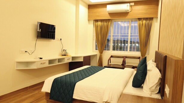 A hotel room with a bed, bedside table, TV, and a window with curtains. - Hotel SKVR Grand - Theni  (Periyakulam)