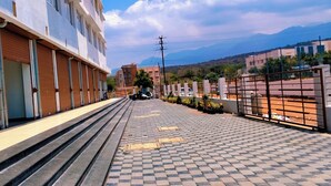 A building with a tiled walkway, a parked scooter, and a view of distant mountains. - Hotel SKVR Grand - Theni  (Periyakulam)