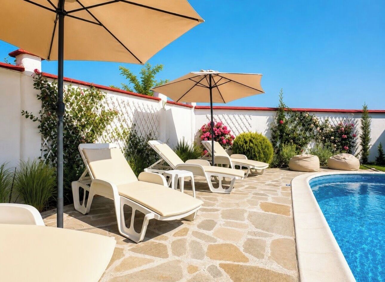 A poolside area with lounge chairs and umbrellas, a stone floor, and a white fence with greenery.