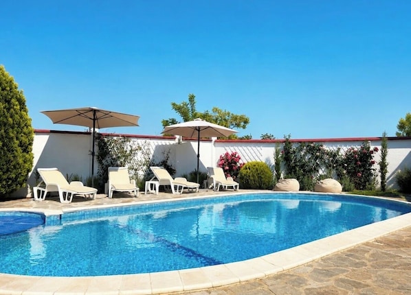 A swimming pool with lounge chairs and umbrellas, surrounded by a white wall and greenery.