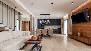 A modern living room with a sofa, a coffee table with chess pieces, a dining area with chairs, a kitchen with a TV, and a staircase. - Villa Krišto ZadarVillas (Privlaka)