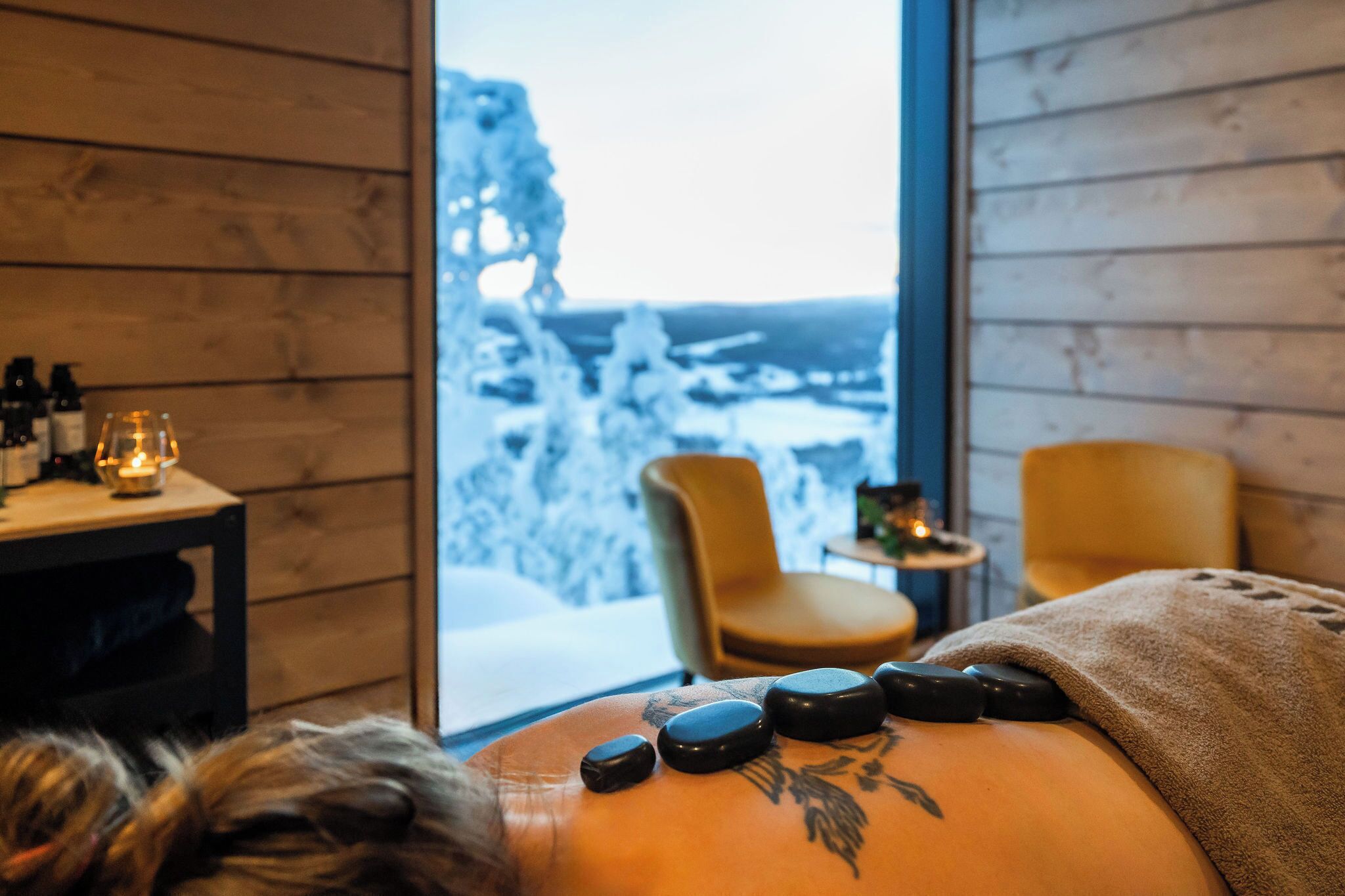 A massage therapy room with a view of a snowy landscape, wooden walls, and a massage table with stones.