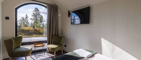 Una habitación con una cama, dos sillas, una mesita, televisor y un ventanal con vistas a árboles y un cielo nublado.
