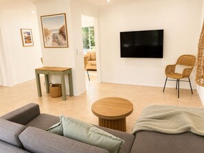 Ein Wohnzimmer mit einer Couch, einem runden Couchtisch, einem Fernseher, einem Korbstuhl und einem Esstisch mit einem Korb.