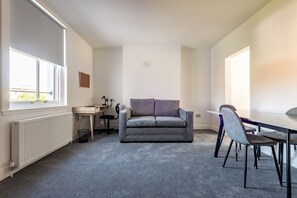 A room with a grey carpet, a grey sofa, a dining table with chairs, a radiator, and a window. - Marine Place | Flat 8 (St Andrews)