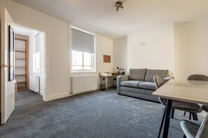 Ein Zimmer mit grauem Teppich, einem grauen Sofa, einem Holzesstisch, einem kleinen Schreibtisch und einem Heizkörper.
