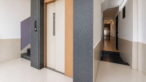 A modern elevator with a wooden panel and a control panel, a hallway with a reflective floor, and a corridor with doors and a light fixture. - FabHotel Metro Residency (Indore)
