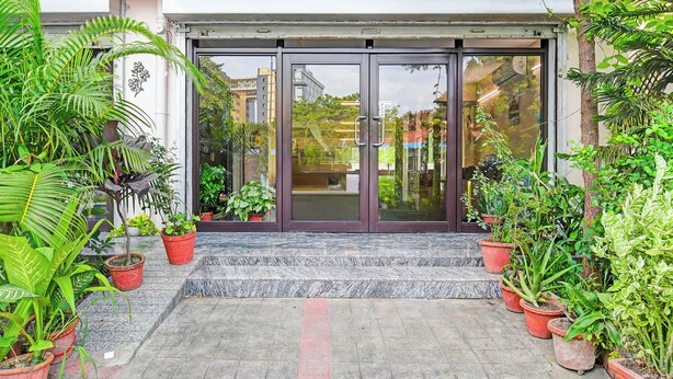 Entrada de un edificio con puertas de vidrio, plantas en macetas y una zona pavimentada.