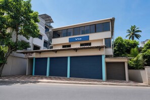 A modern building with a 'Via' sign, multiple garage doors, and a tree-lined street. - Via Nisarg Blue (Alibag)