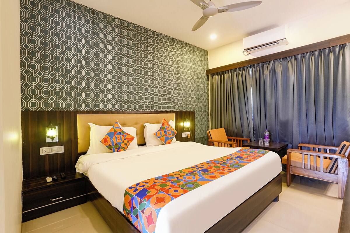 A hotel room with a patterned wallpaper, a bed with a colorful bedspread, a ceiling fan, an air conditioning unit, and a view of the outside through curtains.